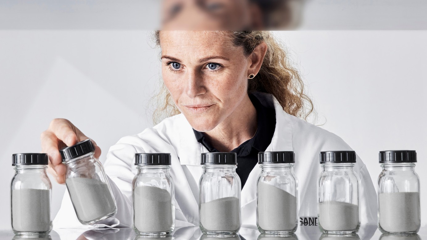 A woman in lab coat behind a table with glass containers with metal powder