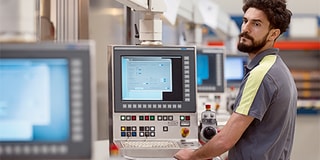 A man in work clothes standing next to a row of monitors and an industrial environment