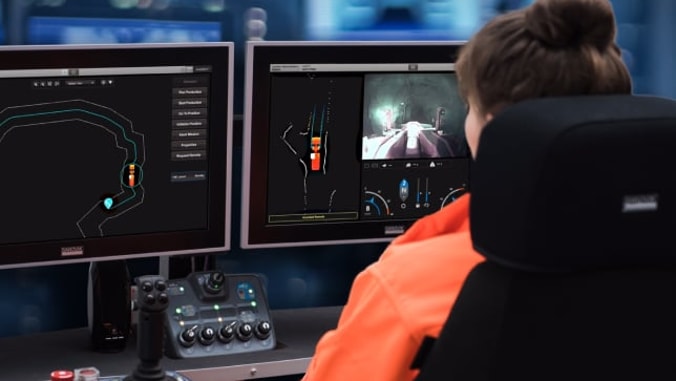 A woman sitting at a desk in a control room looking into two monitors showing different views of a mine