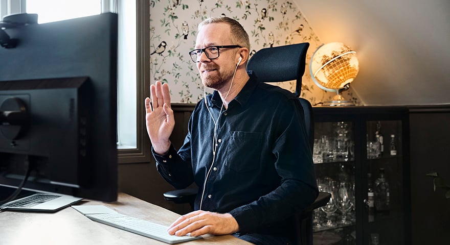 A man with earphones sitting in a home environment at a desk waving towards the computer screen