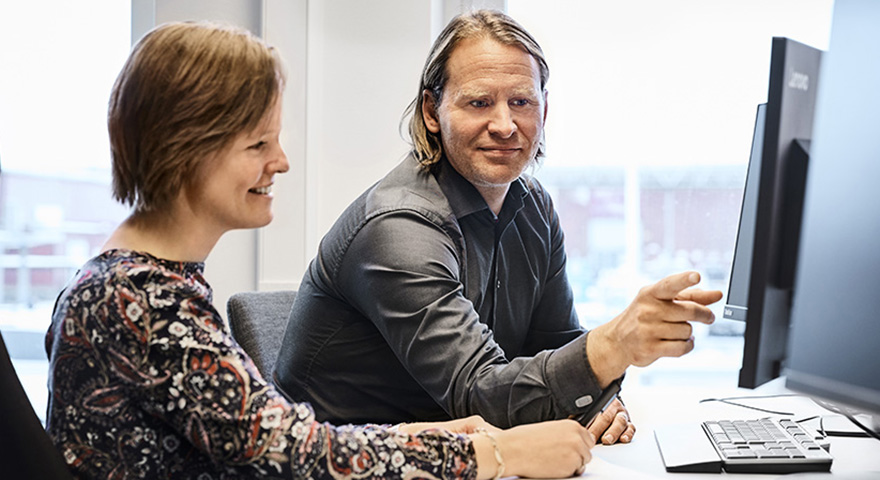 A man and a woman sitting at a desk looking at the same computer and the man is pointing at the screen