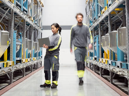A woman holding a laptop and a man walking between two warehouse shelfs