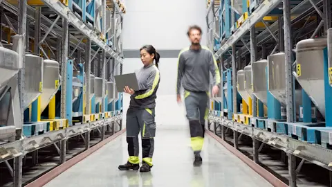 A woman holding a laptop and a man walking between two warehouse shelfs